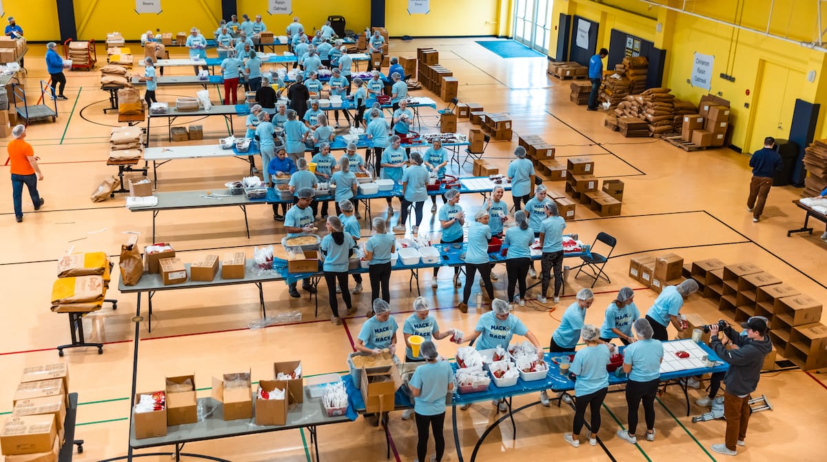 Groups of volunteers circle around tables packing non-perishable meals