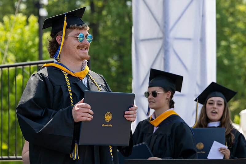 Student wearing round sunglasses holding up ϲʿ diploma at Commencement. 